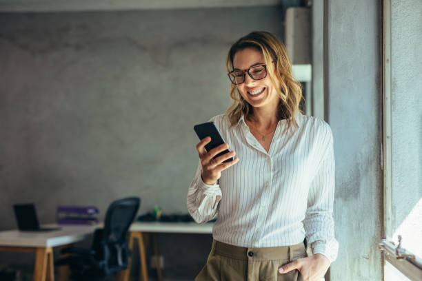 Smiling businesswoman using phone in office Smiling businesswoman using phone in office. Small business entrepreneur looking at her mobile phone and smiling. celular stock pictures, royalty-free photos & images