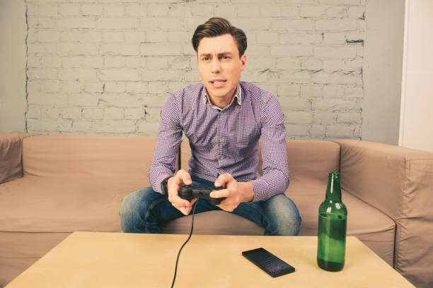 istock Young man sitting on couch and playing video games with beer 1153865516