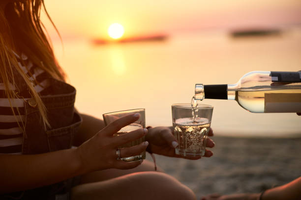 Man hand gracefully pouring white wine while girlfriend holding glasses. Couple enjoy beverage on sunset near sea. Romantic picnic with alcoholic drink on the beach. Friends relax on the shore