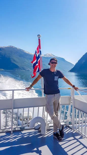 noruega-homem novo que toma um passeio de barco no fjord. bandeira norueguesa de ondulação atrás dele - flam aurlandsfjord sognefjord fjord - fotografias e filmes do acervo