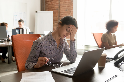 Tired young indian female worker take off glasses feeling unwell suffer from migraine or headache, exhausted sick woman employee touch head have blurry vision or dizziness in shared office