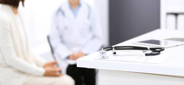 istock Stethoscope, clipboard with medical form lying on hospital reception desk with laptop computer and busy doctor and patient communicating at the background. Medical tools at doctor working table.Medicine concept 1153898435