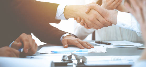 istock Business people shaking hands at meeting while theirs colleagues clapping and applauding. Group of unknown businessmen and women in modern white office. Success teamwork, partnership and handshake concept 1153899297