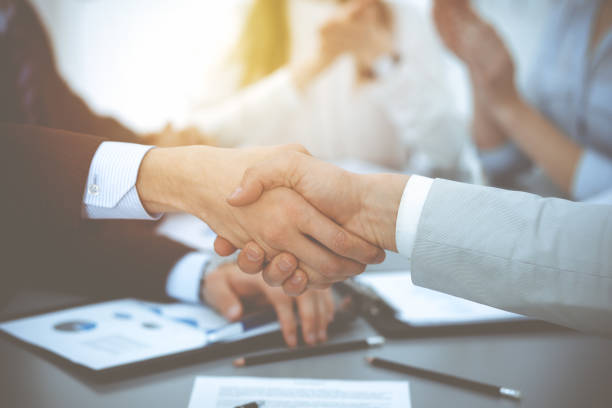 istock Business people shaking hands at meeting while theirs colleagues clapping and applauding. Group of unknown businessmen and women in modern white office. Success teamwork, partnership and handshake concept 1153899301