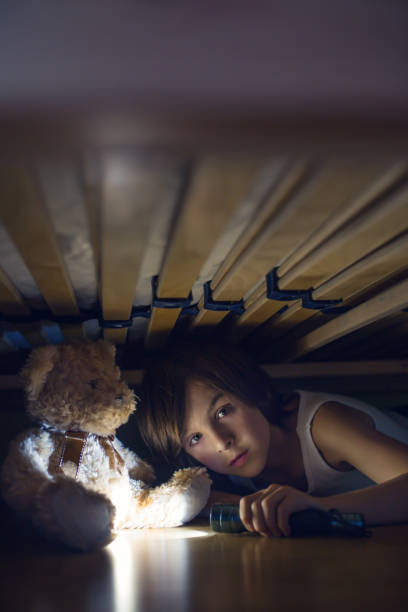 istock Little child, hiding under the bed, hugging teddy bear and holding flashlight, 1153913298