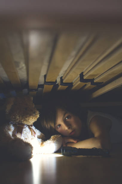istock Little child, hiding under the bed, hugging teddy bear and holding flashlight, 1153913333