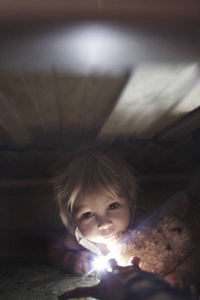 Little child, hiding under the bed, hugging teddy bear and holding flashlight, adult hand trying to reach him