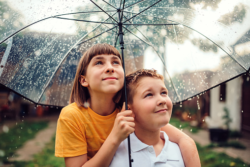 Brother and sister standing underneath clear umbrella and looking the rain falling