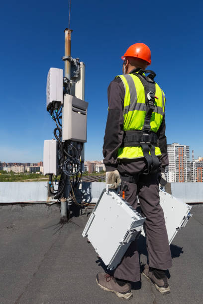istock Telecommunication engineer  in helmet and uniform holds telecomunication equipment in his hand and  antennas of GSM  DCS UMTS LTE bands, outdoor radio units, optic fibers, power cables are installed on the roof. Working process of upgrading telecommunicat 1153920769