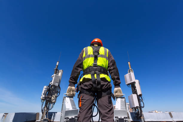 Telecommunication engineer in helmet and uniform holds telecomunication equipment in his hand and antennas of GSM DCS UMTS LTE bands, outdoor radio units, optic fibers, power cables are installed on the roof. Working process of upgrading telecommunication equipment ingeniero de telecomunicación en casco y uniforme sostiene equipos de telecomunicación en su mano y antenas de gsm dcs umts lte bandas, unidades de radio al aire libre, fibras ópticas, cables de alimentación se instalan en el techo. proceso de moderniz