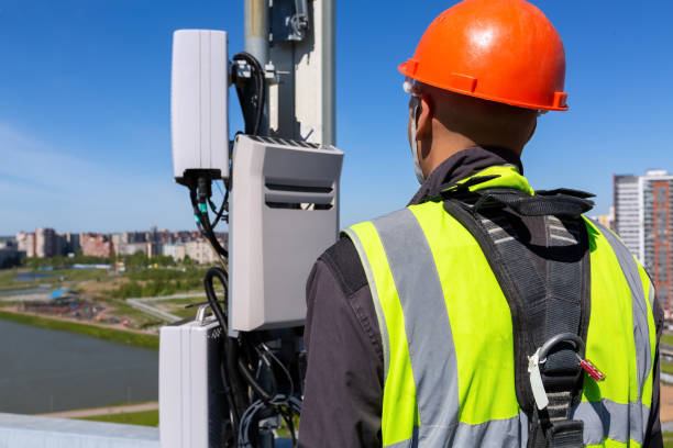 istock Telecommunication engineer  in helmet and uniform holds telecomunication equipment in his hand and  antennas of GSM  DCS UMTS LTE bands, outdoor radio units, optic fibers, power cables are installed on the roof. Working process of upgrading telecommunicat 1153920775