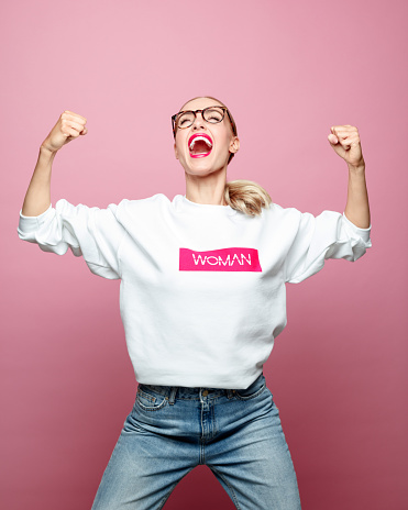 Successful woman clenching fists. Beautiful strong female is screaming against pink background. She is wearing white t-shirt.