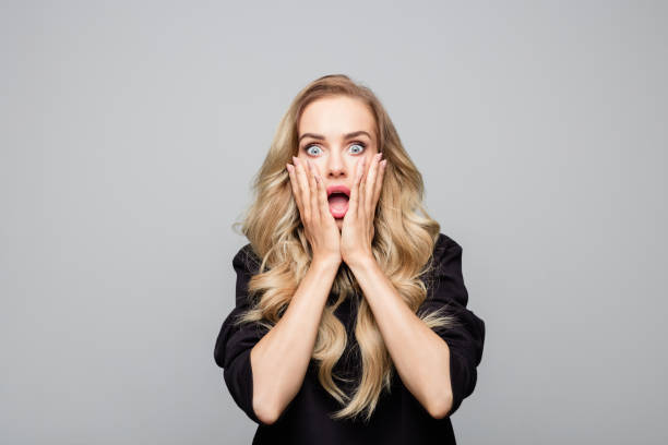 Portrait of shocked young woman with hands on face Portrait of shocked young woman with hands on face. Beautiful blond is wearing black top. She is standing against white background. head in hands stock pictures, royalty-free photos & images