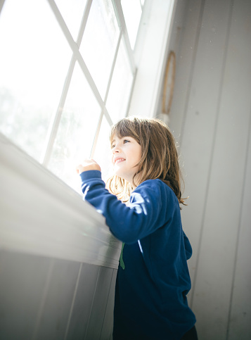 Cute child looks out of a sunny window, the light suggest early morning and she is perhaps wearing pajamas. It looks like a happy positive day. Child looks a bit wistful, Hopeful, unsure, contemplative. A new day.