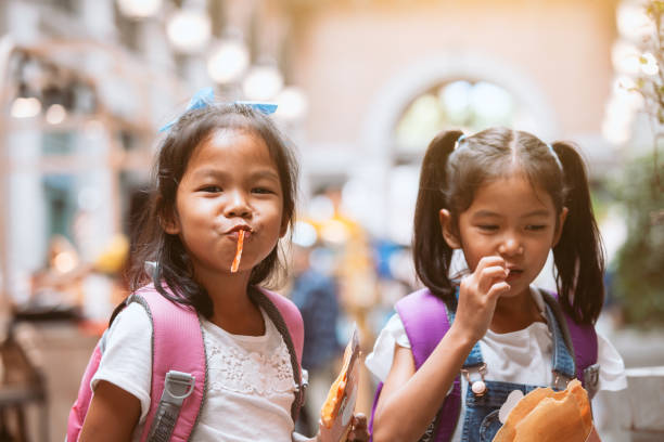 dos lindas niñas asiáticas con mochila comiendo panqueques juntos después de la escuela - eating child cracker asia fotografías e imágenes de stock