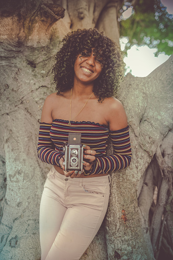 Beautiful young woman in a 60’s-70’s themed image uses a retro old fashioned camera outdoors in a park like location. She is cool and hip and could be from any decade. Photography and vintage retro themed image
