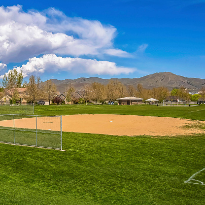 Frame Square Baseball field with view of mountain and cloudy blue sky on a sunny day. Buildings and lush trees can also be seen around the perimeter of the grassy field.
