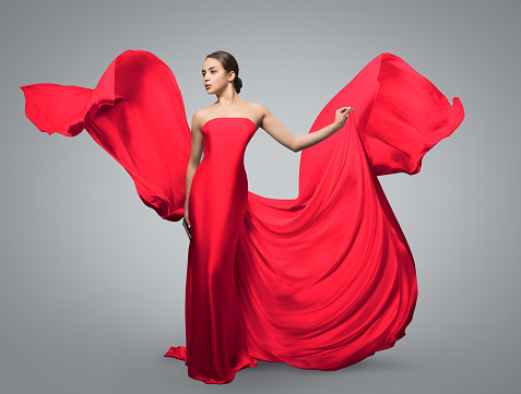 Fashion portrait of beautiful woman in waving red dress. Light fabric flies in the wind. Light gray background. Girl posing studio