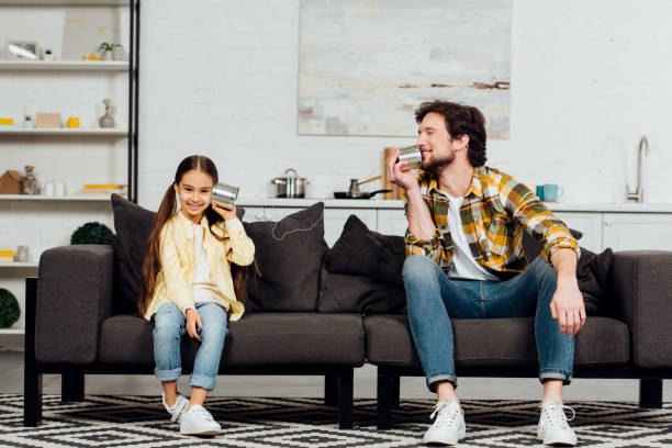 happy daughter holding can phone and cheerful father talking and sitting on sofa happy daughter holding can phone and cheerful father talking and sitting on sofa toy phone stock pictures, royalty-free photos & images