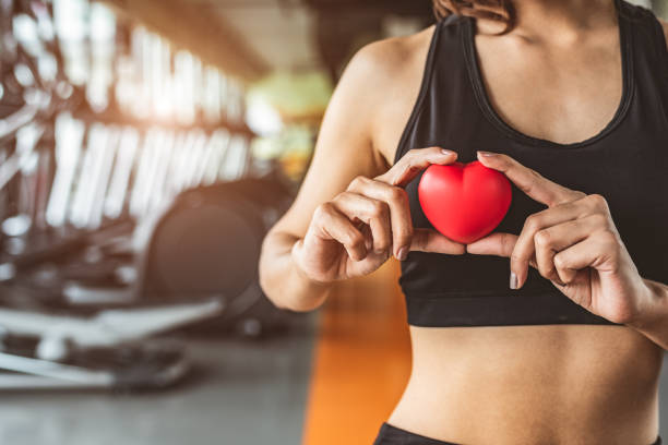 Happy sport woman holding red heart in fitness gym club. Medical cardio heart strength training lifestyle. Pretty female sport girl workout exercise. Cardiac healthy and well-being. Massage ball in ha stock photo