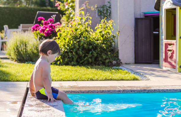 pequeño niño feliz sentado en el lado de la piscina en el jardín jugando con sus pies en el agua divirtiéndose. verano. vacaciones de verano. - swimming pool water people sitting fotografías e imágenes de stock