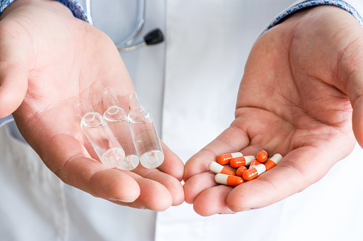 Concept of choice of therapeutic treatment - orange capsules with medication or injectable therapy in form of ampoules with medicinal substance. Doctor holds in one palm ampoules, in another capsules