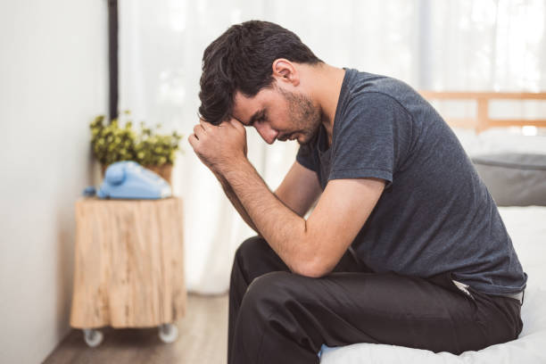Worried man sitting on bed with hand on forehead in bedroom in serious mood emotion. Major Depressive Disorder called MDD concept. Lonely symptom of men alertness. Physical healthcare and social issue stock photo
