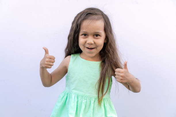 petite fille avec les longs cheveux bouclés bruns d’isolement sur le fond blanc. gosse donnant deux pouces vers le haut - thumbs up human thumb human finger brown hair photos et images de collection