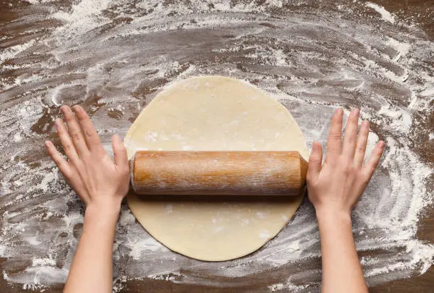 Preparation of pastry. Woman rolling dough on table with flour, top view