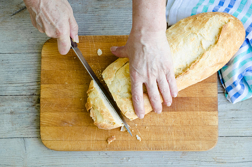 Senior woman cutting freshly baked bread on the wooden board on the rural kitchen table. Traditional bakery concept. Rustic vintage style. Top view.
