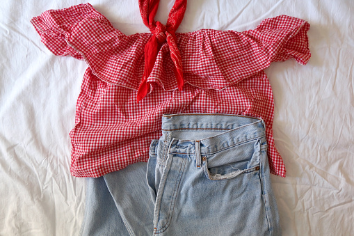 Fashionable outfit with gingham red and white top, worn out jeans and red scarf. Flat lay, white linen background.