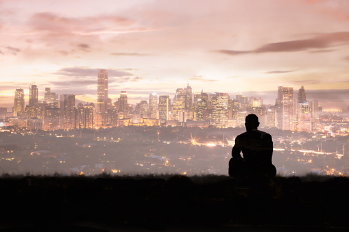 Thoughtful man sitting on a hill looking at the city skyline. Multi exposure image.