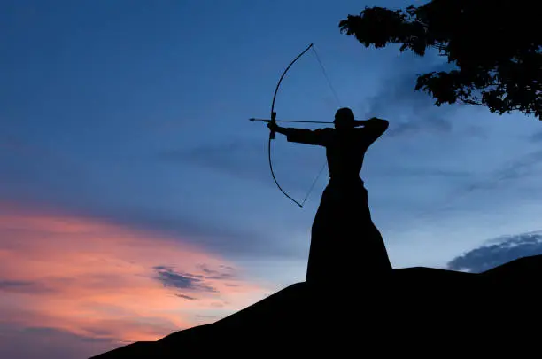 Abstract photo of man silhouette demonstrating martial arts with bow in front of sunset sky