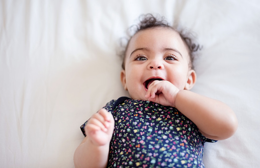 Shot of an adorable baby girl lying on bed with hand in mouth looking at camera and smiling