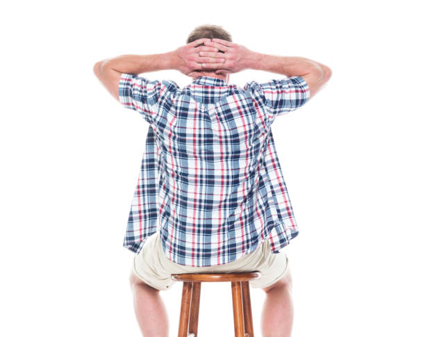 istock Handsome man in shirt and shorts is sitting on stool, rear view 1154154746