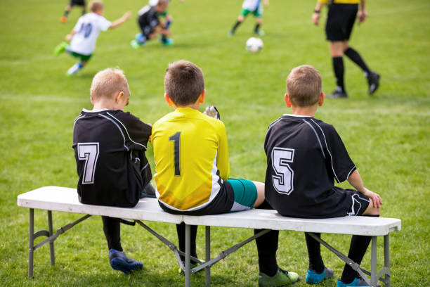 ławka piłkarskia. młodzi piłkarze siedzący na ławce rezerwowych piłki nożnej. mecz piłki nożnej i sędzia w tle - soccer bench soccer player sport zdjęcia i obrazy z banku zdjęć