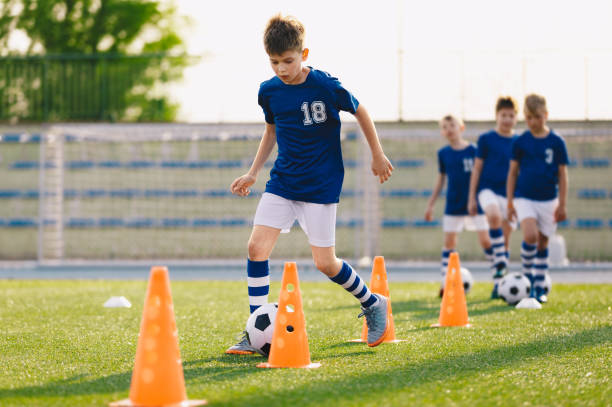 football drills: der slalom-bohrer. jugendfußball-übungsübungen. junge fußballer trainieren auf dem platz. fußball-slalom-kegelbohrer. junge in blauen fußball-trikot-shirt läuft mit ball zwischen kegeln - mannschaftsfußball stock-fotos und bilder