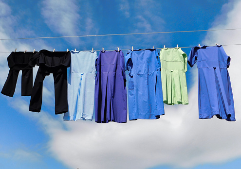 Handmade Amish dresses and black pants drying on a clothing line in the breeze, with blue skies and clouds in the background