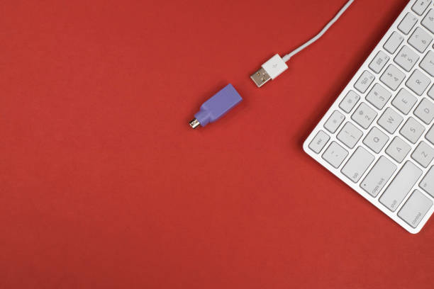 A wired Apple keyboard with a PS2 adapter to plug into older computers.  There is copy space for print.  The background is a single, vibrant, saturated color (red).  The keyboard is silver and white.  This can represent a clean, simple and tidy desktop for work.