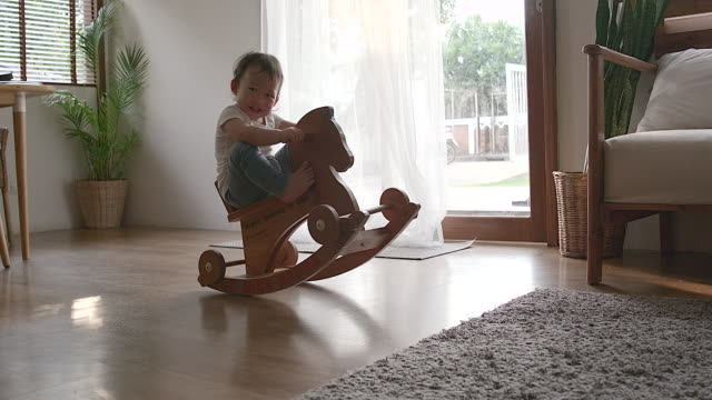 Funny babysitting on the toy horse.Full-length portrait of a little lovely smiling girl wearing a white shirt and blue pants swaying on the wooden toy horse.