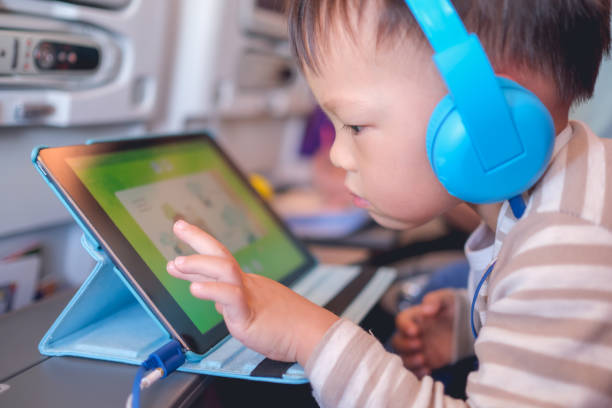Asian 2 - 3 years old toddler boy child wearing headphones using tablet pc watching cartoons / playing game during flight on airplane stock photo