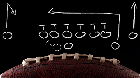 American football tactics scheme, sports coaching and offensive play concept with a ball in front of a blackboard with a game plan drawing done by the coach with dramatic lights