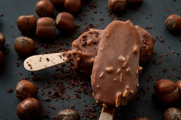 Frozen dessert and summer snack concept with close up on two delicious chocolate and hazelnut ice cream bars surrounded by scattered hazelnuts, cocoa powder and shaved chocolate on dark background