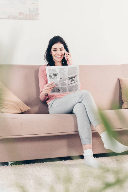 istock selective focus of beautiful smiling woman sitting on couch, reading newspaper and using smartphone at home 1154319730