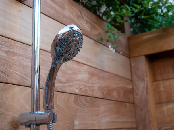 Handheld steel showerhead hanging outside within a wooden fence area and trees