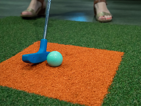 Blue putter on turf next to green golf ball on miniature golf course with a woman hitting