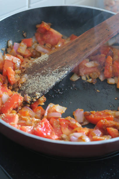 Image of fresh chopped tomatoes and diced red onions being stir fried in hot non-stick frying pan with wooden spatula spoon on hot kitchen cooker hob, tomato and onion base for ratatouille or spaghetti Bolognese pasta sauce, browning and caramelising stock photo