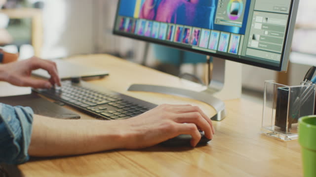 Close Up Shot of Designer's Hands Working in Image Editing Software on His Personal Computer with Big Display. He Works in Cool Office Loft. Tablet and Headphones Lie on the Table.
