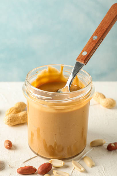 istock Glass jar with creamy peanut butter and spoon, and peanut on white table against light background, space for text 1154353108