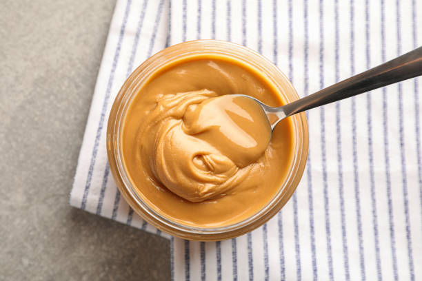 istock Glass jar with peanut butter and spoon, and kitchen towel on grey background, space for text and top view 1154355528
